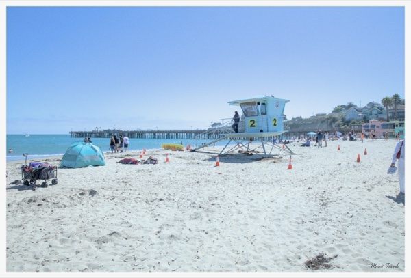 Beachgoers gather around a lifeguard tower with a pier in the distance.